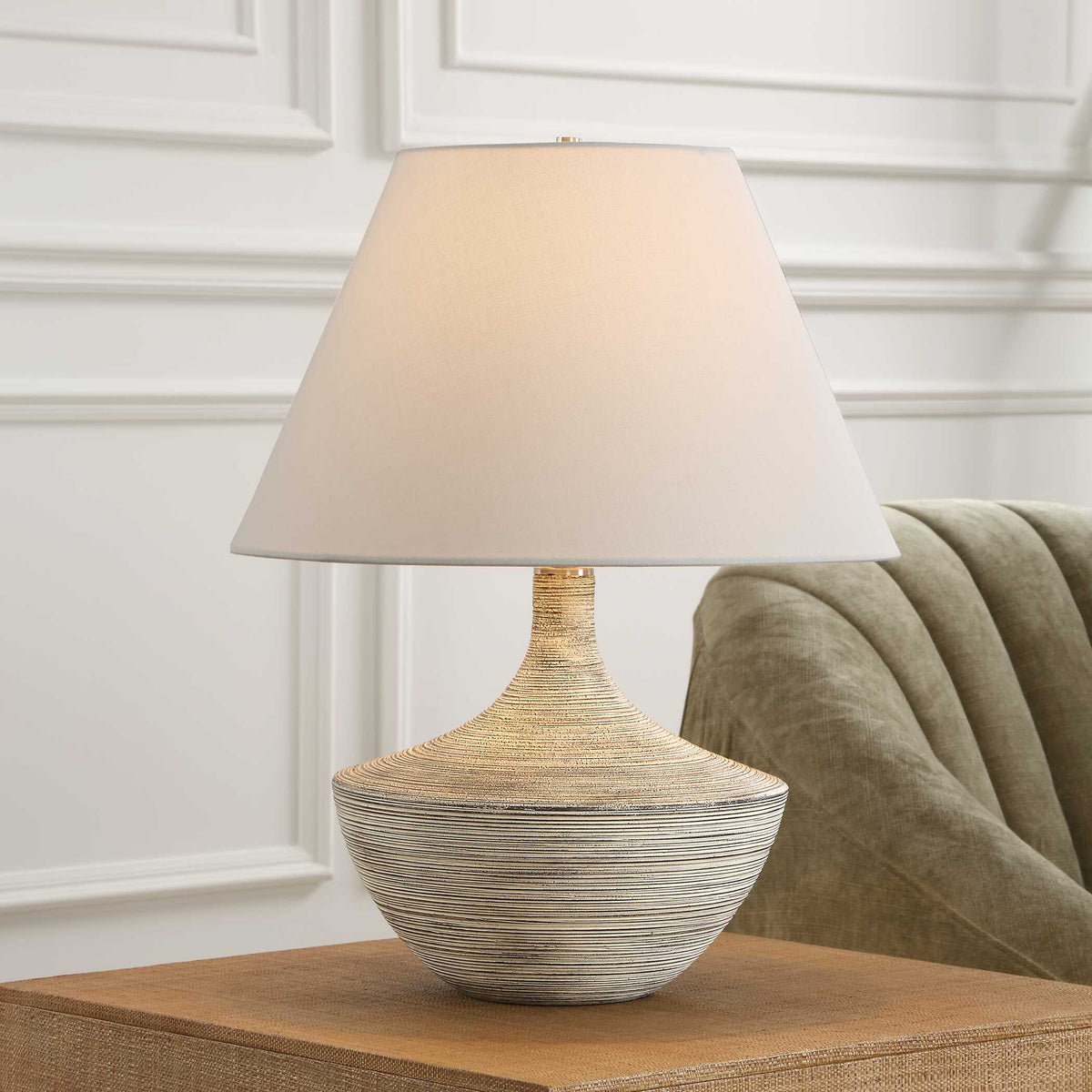 Carafe ribbed ceramic table lamp on a woven console beside a sage green channel tufted sofa