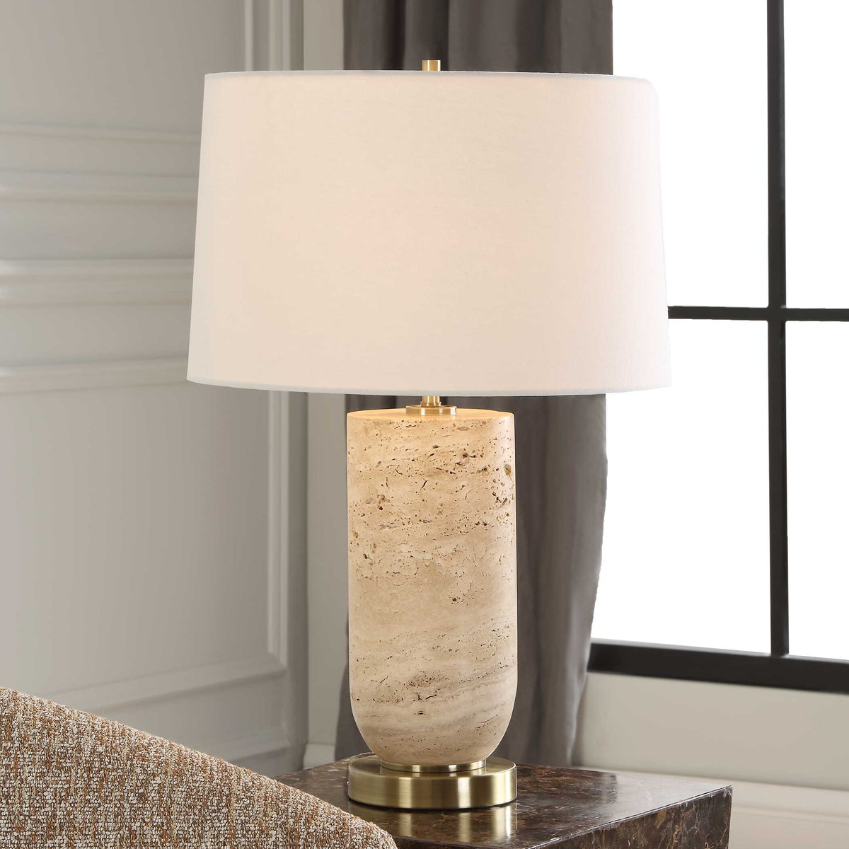 Aubrey travertine table lamp lit on a dark marble side table beside a tweed armchair and window