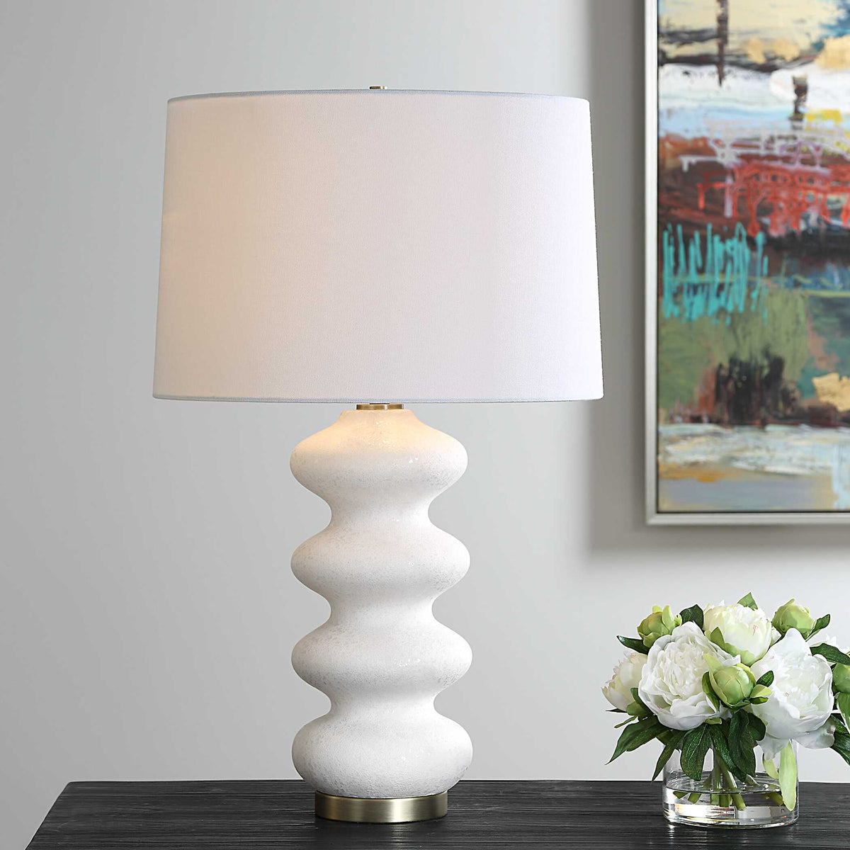 Liora white porcelain table lamp lit on a dark wood table beside white peonies in a glass vase