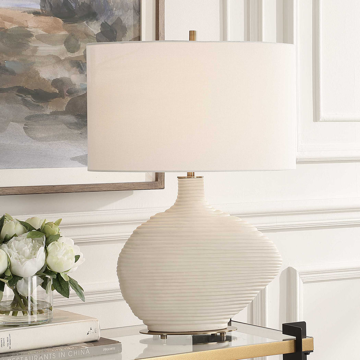 Duostacked white ceramic table lamp with oval shade on a glass and gold console beside peonies and books