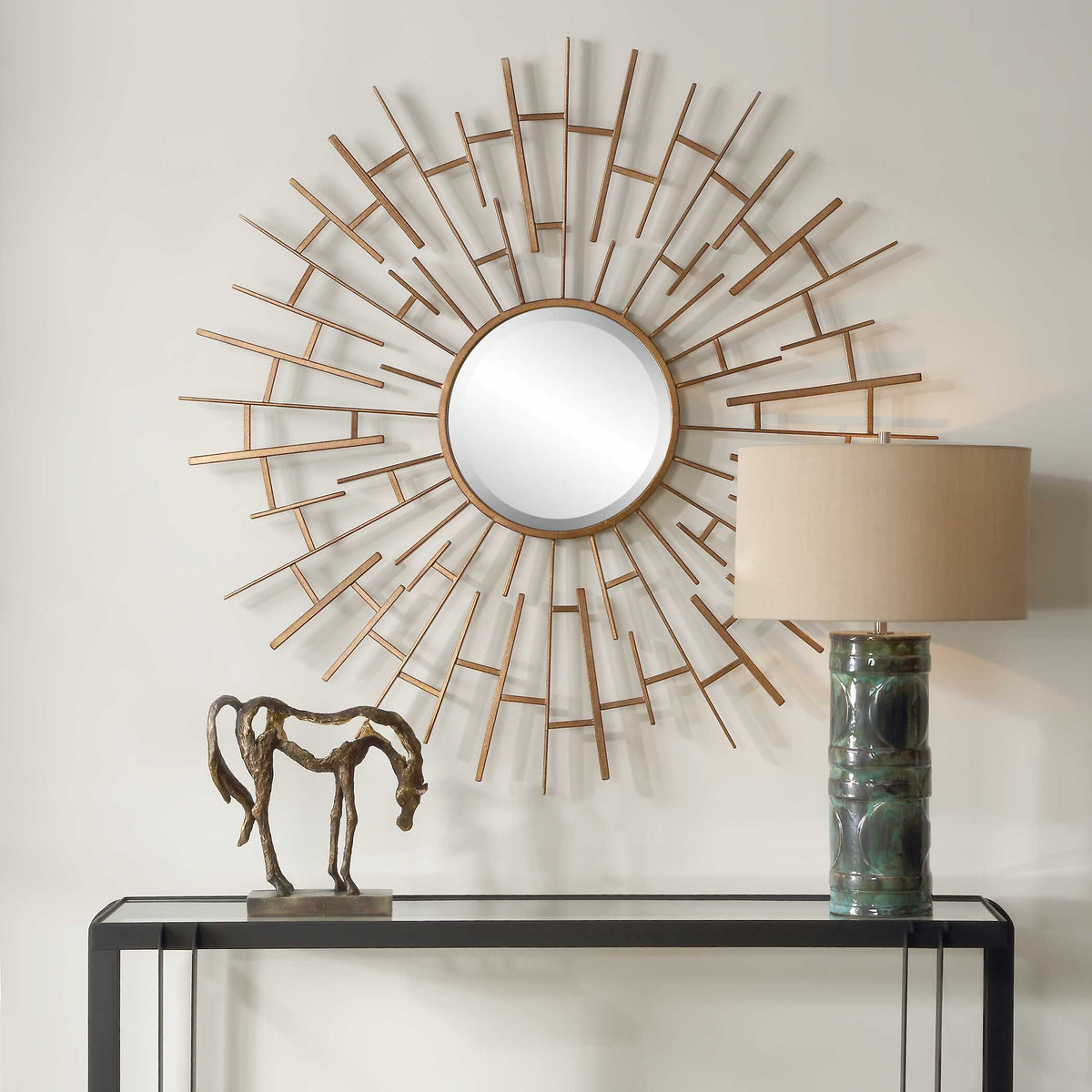 Round sunburst wall mirror with antiqued gold iron frame above a console with a table lamp