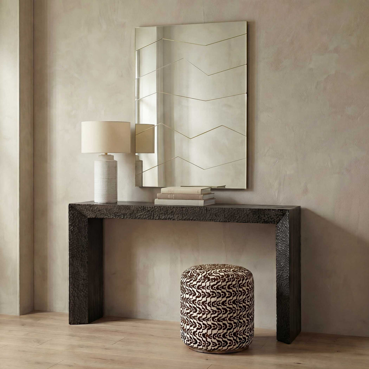Faceted gold mirror over a textured dark console table with a white lamp and patterned pouf