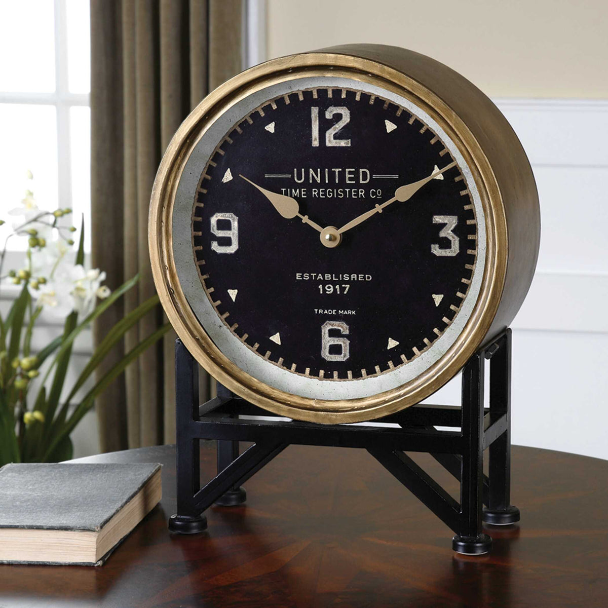 Black and gold table clock on a dark wood table beside a book and orchids