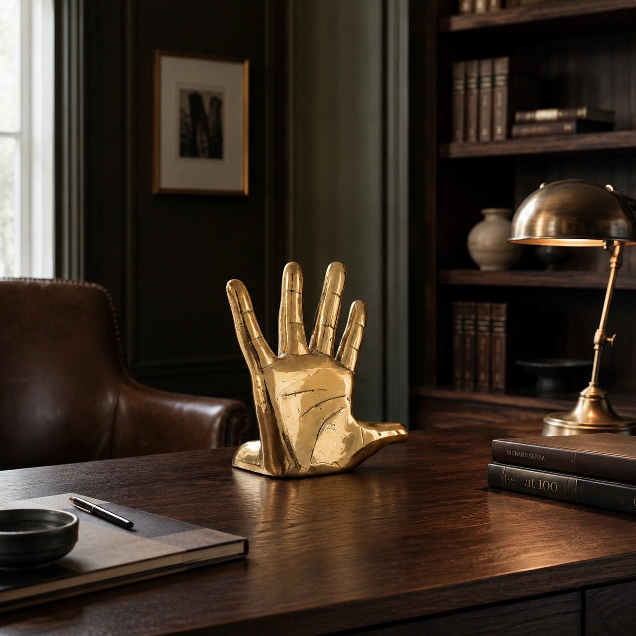 Gold hand sculpture on a wooden desk with books and a lamp in a room setting.