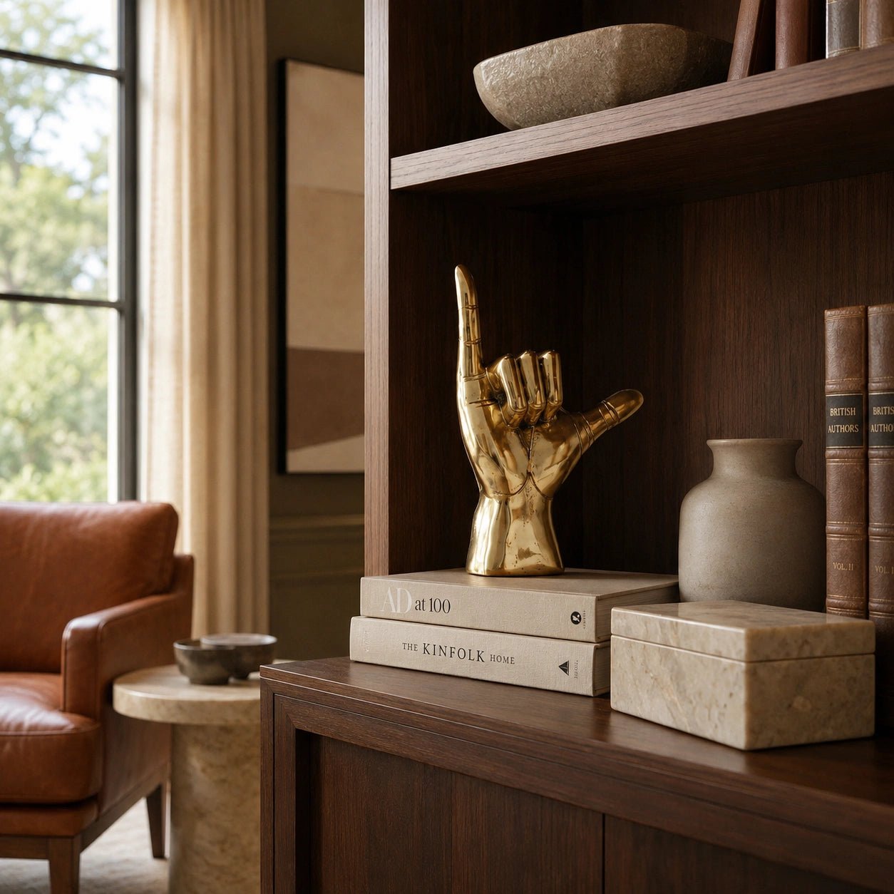 Decorative gold hand sculpture on books on a wooden shelf with a brown armchair and window in the background.