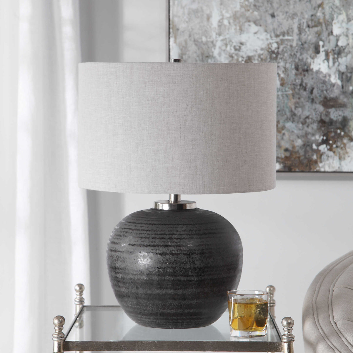Gray round ceramic bedside lamp with brushed nickel hardware in front of sheer curtains and a gray painting
