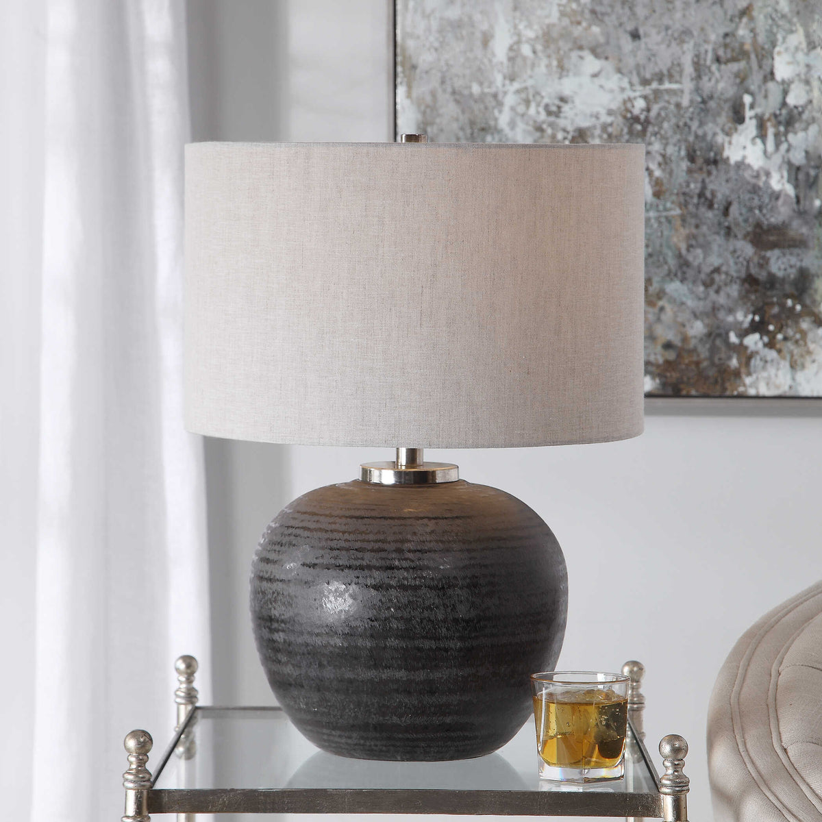 Mikkel gray textured ceramic table lamp on a mirrored side table beside a glass of whiskey and abstract art