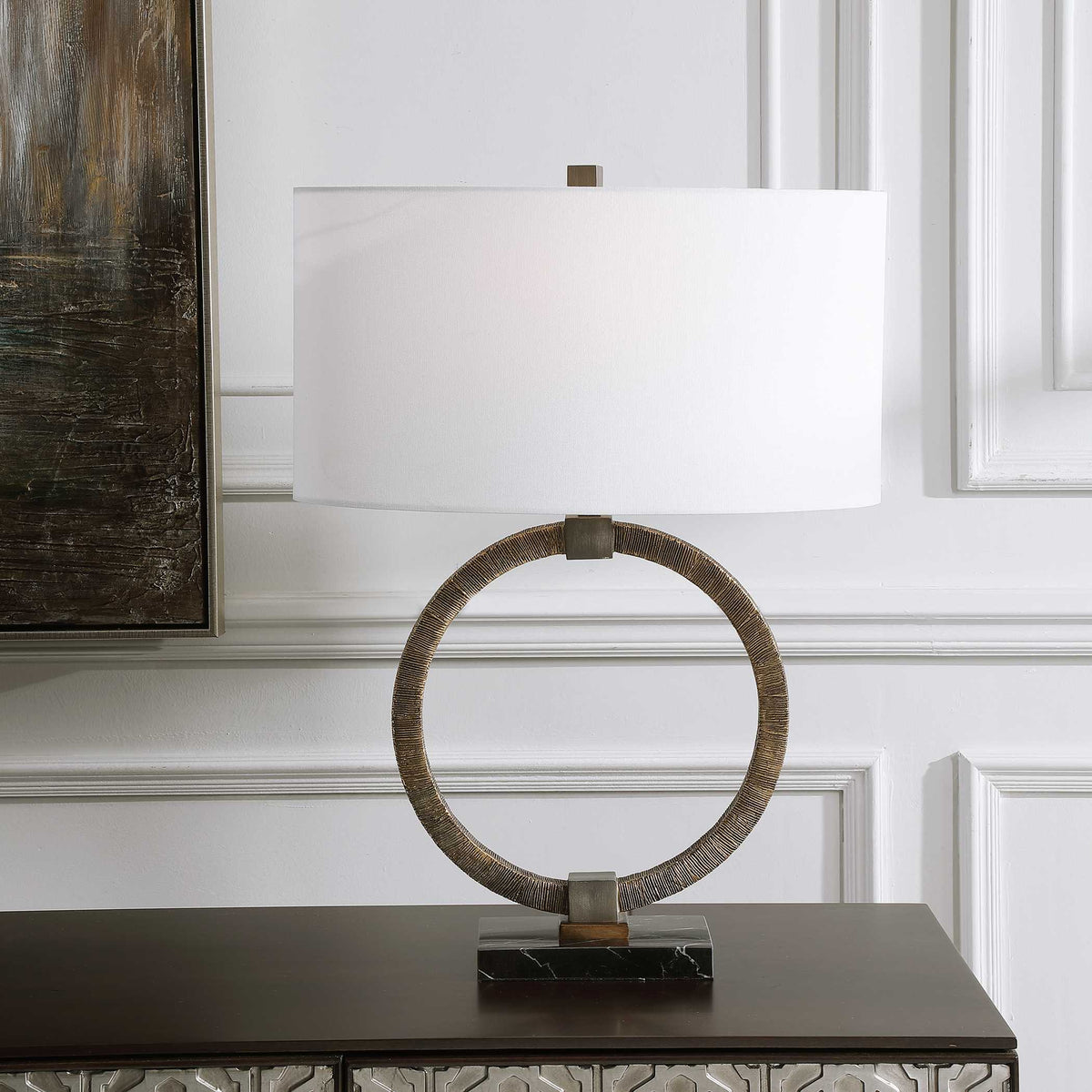 Relic antique gold open ring table lamp on a dark wood console against white wall paneling and framed art