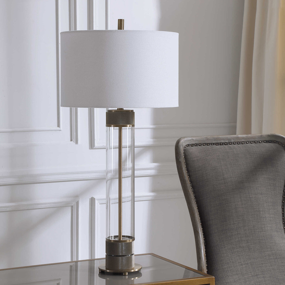 Clear glass column bedside lamp, unlit, on a brass frame side table against white wall paneling