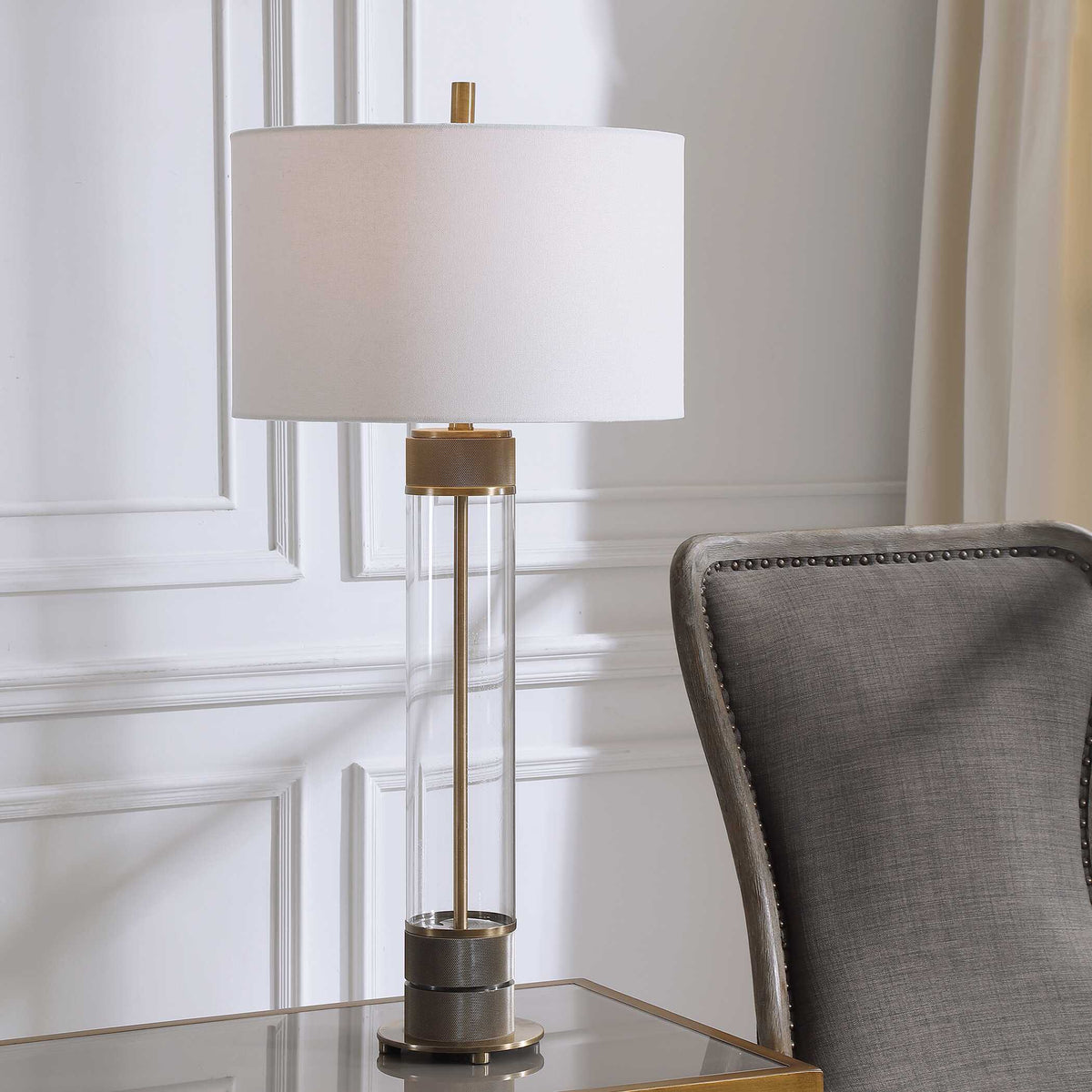 Anmer clear glass table lamp lit on a glass top side table beside a gray wingback chair with nailhead trim