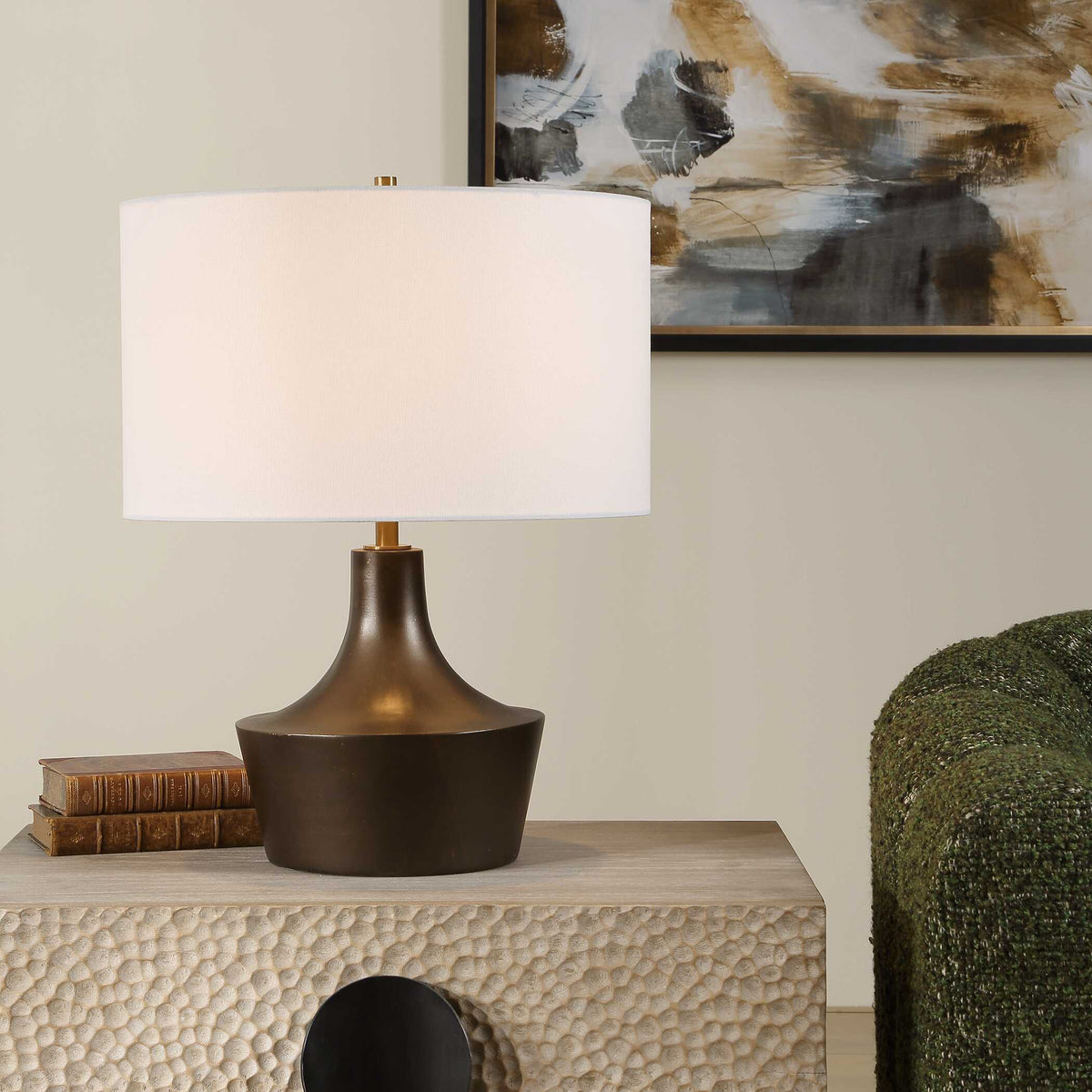Paseo bronze curved table lamp on a hammered cream side table beside stacked books and a green armchair