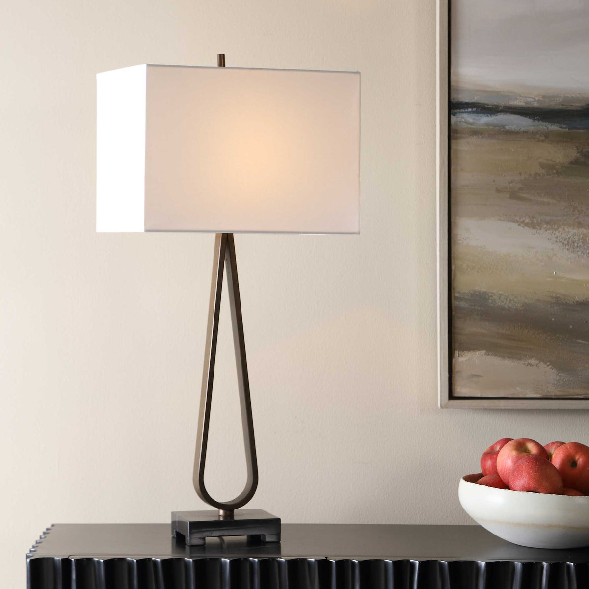 Galloway slim dark bronze loop table lamp on a fluted dark console beside a bowl of apples and landscape art