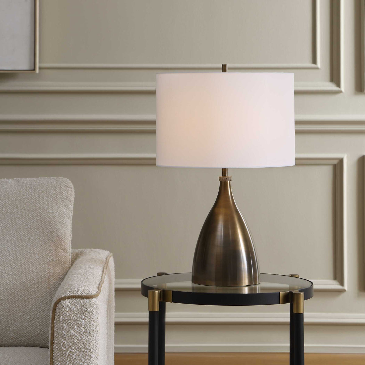Jereko bronze table lamp on a round glass side table beside a boucle armchair in a paneled room