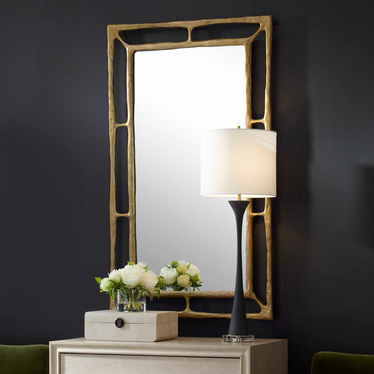 Brass accent mirror above a light cabinet with a table lamp on a dark painted wall