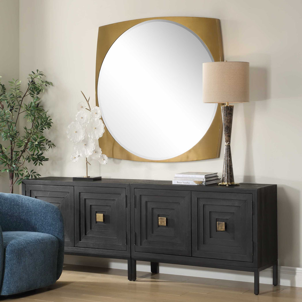 Round glass in a square brass frame over a credenza beside a blue accent chair, angled view