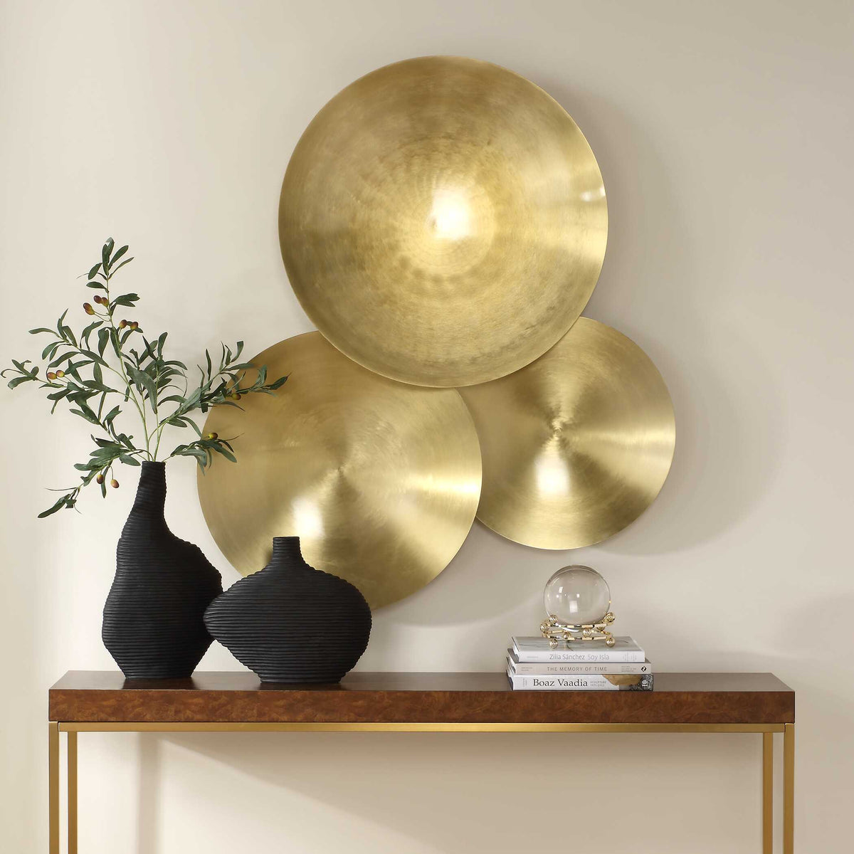Cymbalic metal wall decor above a wood and brass console with black vases and olive branches