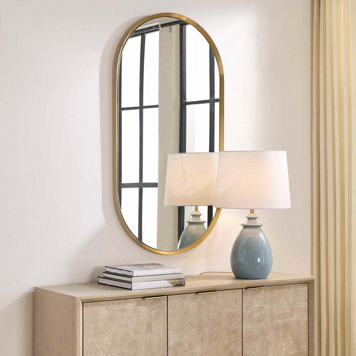 Brass oval vanity mirror over a credenza reflecting a window, angled view