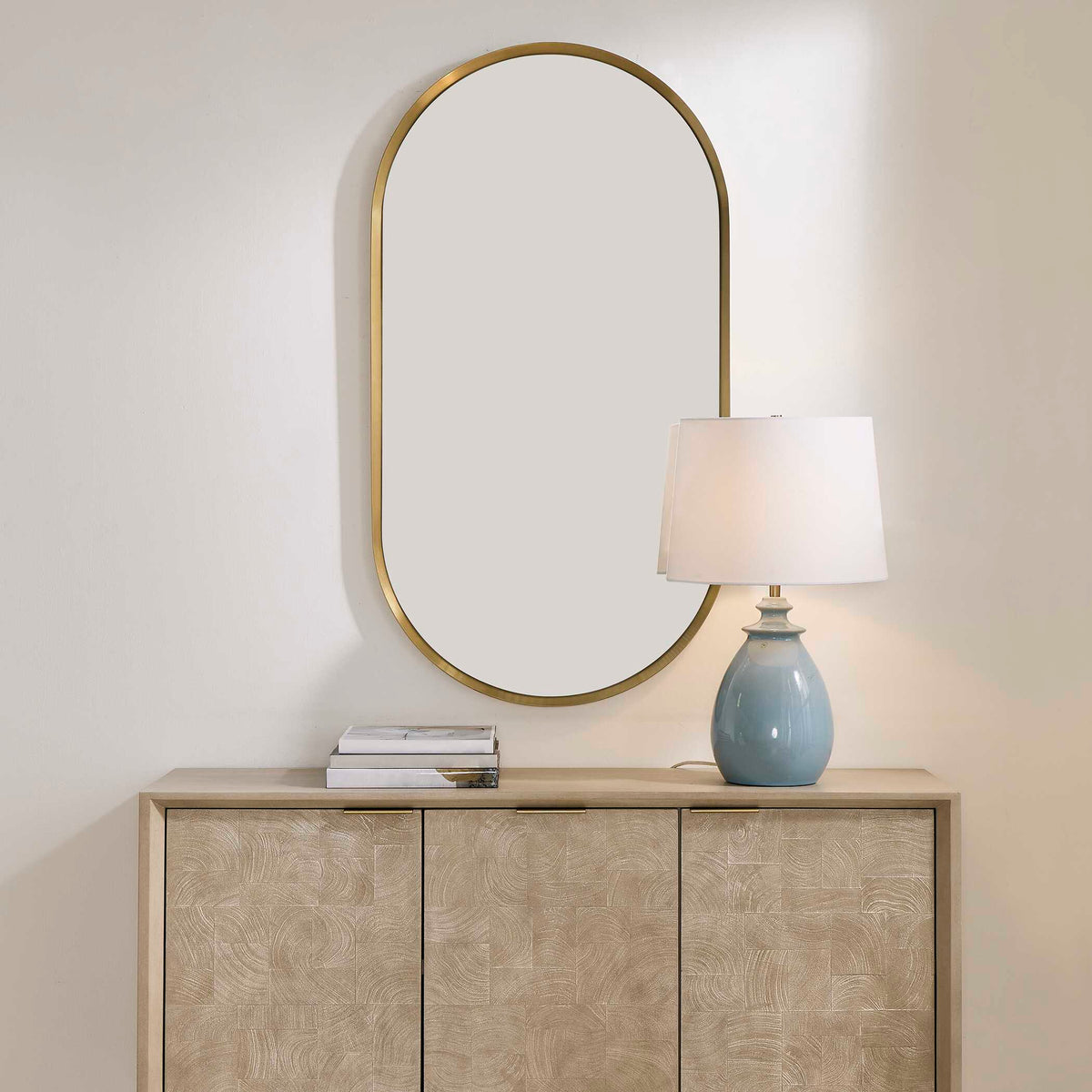 Uttermost 08298 Torrado collection oval mirror above a light cabinet with a blue table lamp