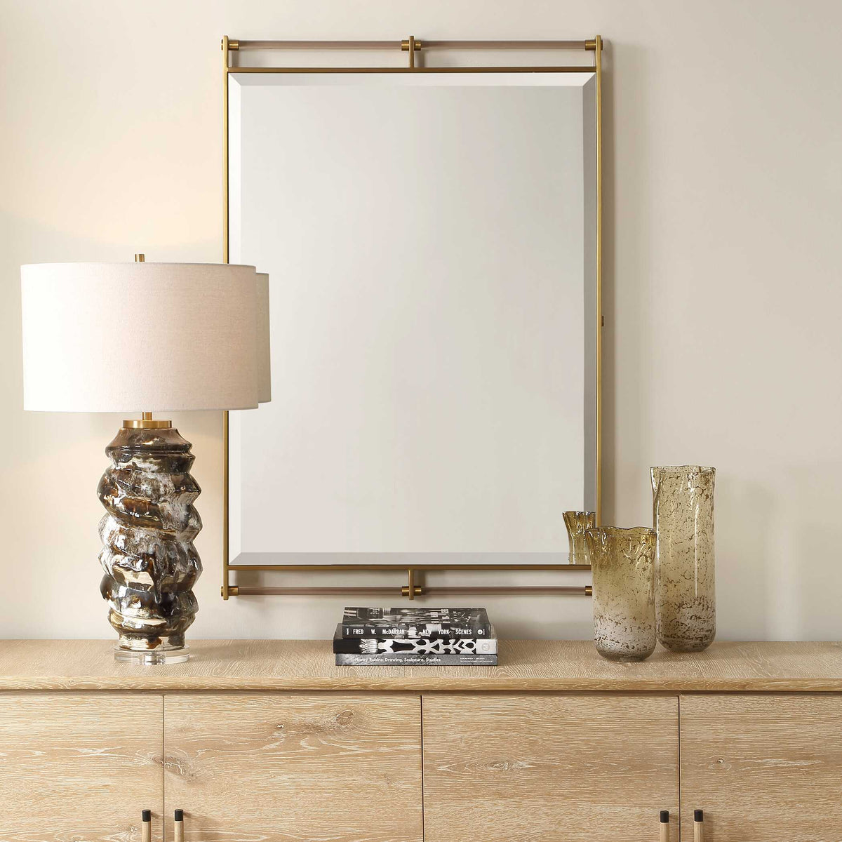 Uttermost 08318 Ripoll collection mirror above a light wood credenza with a glass table lamp and amber vases