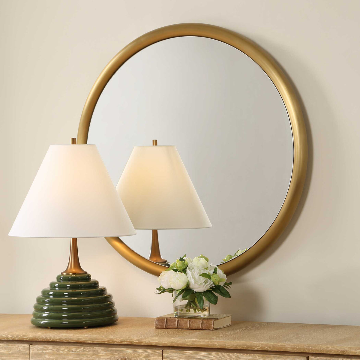 Circle mirror with a smooth brass ring frame, angled behind a lamp and white flowers