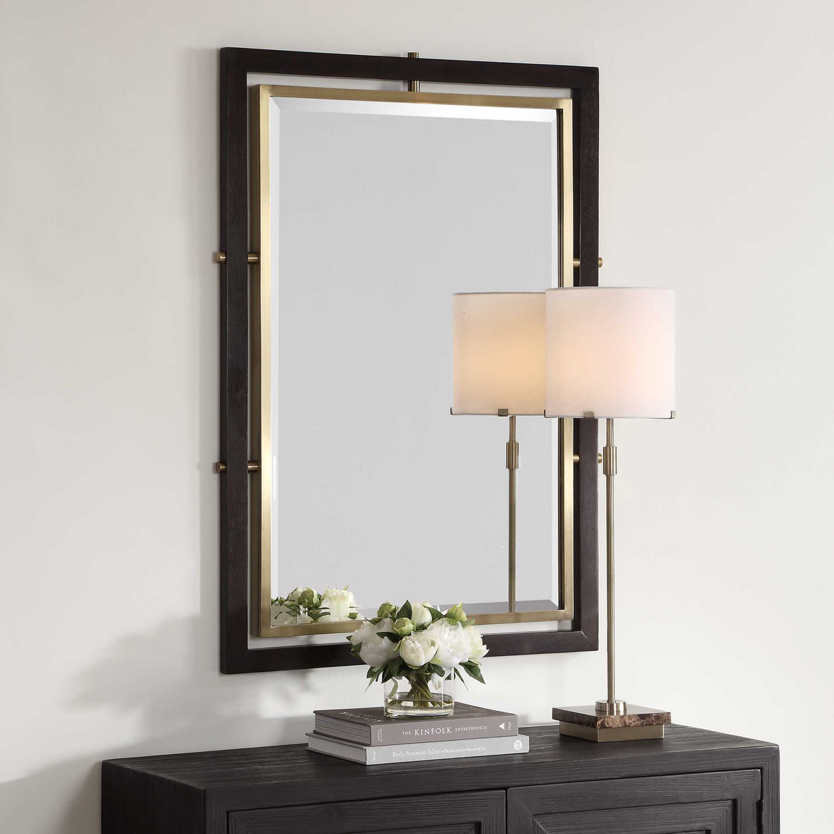 Two tone wall mirror above a dark cabinet with a table lamp and white flowers