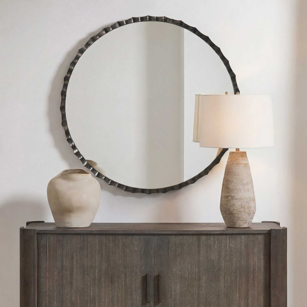 Uttermost 08415 Grander round mirror above a dark wood cabinet with a textured lamp and stoneware vase